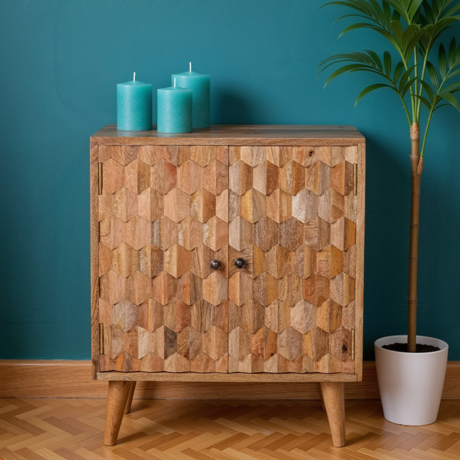 Wooden cabinet with geometric carved doors, two teal candles on top, teal wall and potted plant.